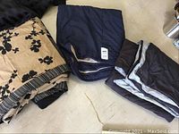 Three folded quilts: one queen quilt with beige background and black floral print, two light wrap quilts in dark solid colors (navy and brown) with some trim details.