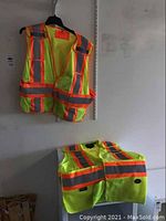 Two high visibility safety vests hanging on a wall, showing front and back reflective striping.