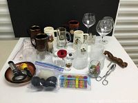 Full view of all bar glassware, mugs, drink markers, and utensils including mugs with brewery and holiday print, textured glasses, stemmed glasses, corkscrew, tongs, bottle opener, and accessories in bags.