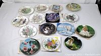 Photo showing 15 decorative plates laid out on white surface, various floral and nature-themed artwork, and child portraits in the lot.