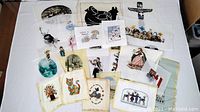 Photo showing a collection of 18 needle point artworks laid out on a white surface with various subjects including animals, people, and scenic images.