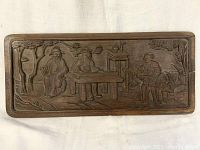 Full front view of the rectangular antique carved wooden panel showing four Asian figures around a table outdoors with trees and rocks.