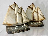 Pair of cast iron Blue Nose ship bookends showing full front view, painted with weathered white sails, black bases with wave texture and inscription 'BLUE NOSE'.