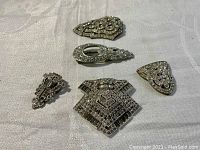 Five antique decorative metal pieces with detailed rhinestones on a fabric background. Variation in shapes and intricate patterns visible.