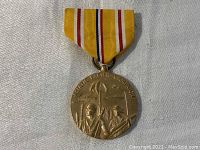 Front view of the Asiatic Pacific Campaign WWII pin with two soldiers and a flag in bas relief and the inscription 'Asiatic Pacific Campaign'. The pin is attached to a yellow ribbon with red, white, and blue stripes.