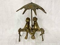 Front view of bronze sculpture showing two frogs seated together under a textured umbrella, on a bench with decorative legs.