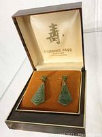 Pair of green jade drop earrings in brown hinged jewelry box with orange velvet cushion and satin-lined lid with gold Chinese character and branding.