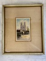 Full view of framed hand-colorized etching of Paris Notre Dame with surrounding beige mat and thin frame showing wear.