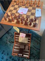 Well-made wooden chess set with light and dark pieces set up on a folding wooden chessboard table. Handwritten 'For Sale' and lot tags visible.