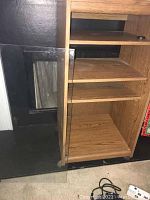 Wood grain stereo cabinet with the glass door off the hinges, door leaning nearby on the floor.