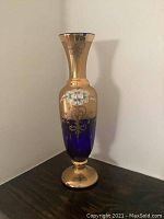 Front view of tall blue glass vase showing gold gilded neck and base with hand painted white floral decoration.