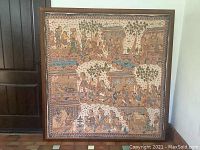 Full view of the batik framed artwork showing detailed human figures in traditional Indonesian clothing, trees, and houses in a horizontal rectangular wooden frame.