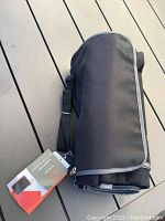 Rolled up black picnic blanket with attached black tote and strap, tag visible showing brand.