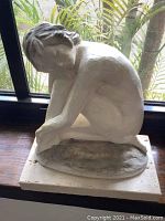 Front view of the white sculpture of a seated pensive lady on the base, in front of a window with natural light.