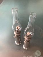 Two silver and glass oil lamps standing next to each other on a smooth surface, showing full height and base.
