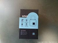 Front view of the Smart Loq Biometric padlock box showing icons of fingerprint scanner, waterproof rating, app connectivity, and what the box contains.