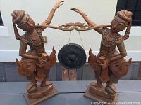 Full view of two connected hand-carved wooden figures with a hanging gong between them on a wooden bar.