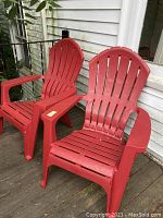 Two red Adirondack chairs on a wooden deck beside the exterior wall of a house, showing front and side views.