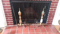 Full view of brass andirons, poker, and wire log holder placed in front of fireplace.