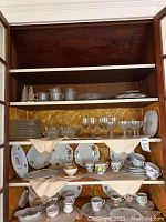 Wooden shelf with assorted china teacups, saucers, plates, and glassware items arranged on three levels, all with floral and monogrammed patterns mostly in white porcelain.