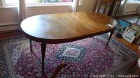 Oval wooden dining table in natural wood tone with four turned legs and attached leaves removed, placed indoors on a patterned rug.