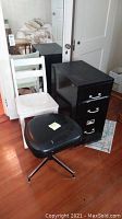 Image showing black steel file cabinet with white painted wood chair and black backless office stool on wooden floor.