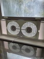 White dual-blade window fan installed in window frame, showing front view of fan blades and surrounding frame
