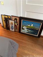 Five framed artwork pieces leaning against wall on wooden floor. Includes needlepoint inspired portraits and lighthouse painting. One frame shows paint stain.