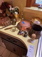 Overview of home decor items arranged on countertop: copper pan, brass bell, brass tall vase with flowers, brass container, copper pot, decorative bowls, figurines, and candy dish.