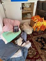 Pile of assorted linens and textiles including folded pink, green, and blue gray fabrics, orange knitted throw, stored on floor and baskets