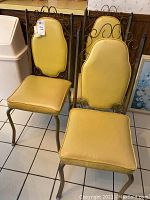 Front angled view of two MCM metal and vinyl side chairs showing the yellow vinyl cushions and metal backs with scroll design.