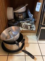 Photo showing Cuisinart hand mixer, cake pans, metal cookware, plastic bowls and kitchen equipment stacked inside a wooden cabinet and on floor.