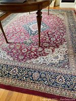 Photo showing the rug under a table, highlighting the red field with beige and blue floral motifs and central medallion.
