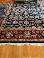 View of most of the rug showing floral pattern on black background with red and beige border and fringes on one end, placed on a wooden floor.