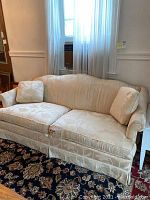 Full front view of a three-seat upholstered sofa in light beige floral fabric, showing wear, staining and fabric damage. Two matching cushions are placed on the sofa.