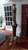 Full view of gold-tone Greek-style female statue on the tall ornate pedestal with other items around base