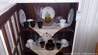 Shelf displaying white floral teacup and saucer sets, dark blue or black gold leaf Greek patterned teacups and saucers, clear tall glass vase, amber and clear glass bowls, floral pattern plates.