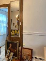 Wall mirror with wood frame hanging on wall, visible hooks, and three framed artworks leaning against wall and each other on the floor.