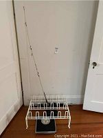 Fishing rod resting in a white metal shoe rack with a mechanical floor scale beneath it, positioned against a beige wall.