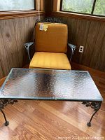 Wrought iron chair with mustard cushions alongside wrought iron coffee table with glass top in a wooden-paneled room.