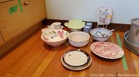 Multiple Asian spoons, bowls including scalloped and floral, plates stacked, white porcelain tray, decorative plates set on wooden floor.