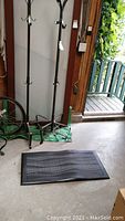 Two tall black metal coat racks standing upright on a green tarp on the floor, and a black cushioned floor mat laid out in front of them on a concrete floor, all indoors near a doorway.