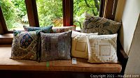 Seven various decorative cushions placed on a bench by a window. Includes embroidered, patchwork, and neutral tone cushions, largest 19x19 inches. One cushion shows wear on the corner.
