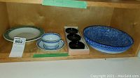 Shelf showing decorative pottery plate, blue and white teacup with saucer, six black lacquer coasters in box, and a blue textured porcelain bowl.