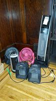 Photo showing three heaters and three fans on a wooden floor with dark wood paneling behind. Includes small black heater, pink fan, black small fan, tall silver Honeywell heater, and another black heater.