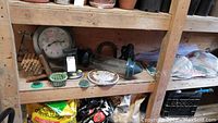Wide view of wooden shelving with outdoor clock, hummingbird feeder, flower frogs, plate covers, cushions, and miscellaneous garden accessories