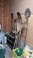 Wide view of the garden tools and equipment grouped together against wood plank wall showing rakes, shovels, sprayer, shelf, and green container.