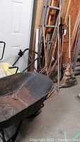 Overall lot showing metal wheelbarrow with black pneumatic tires and garden tools against a wall including rakes and broom.