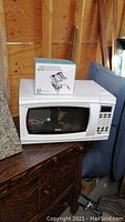 White RCA microwave oven model RMW1112 with Arcosteel pasta maker box on top, placed on dark wooden furniture in unfinished wooden room.