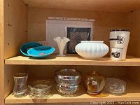 Two wooden shelves displaying various items including glass dishes, pottery, decorative plates, milk glass vase, and acrylic paintings in the background.